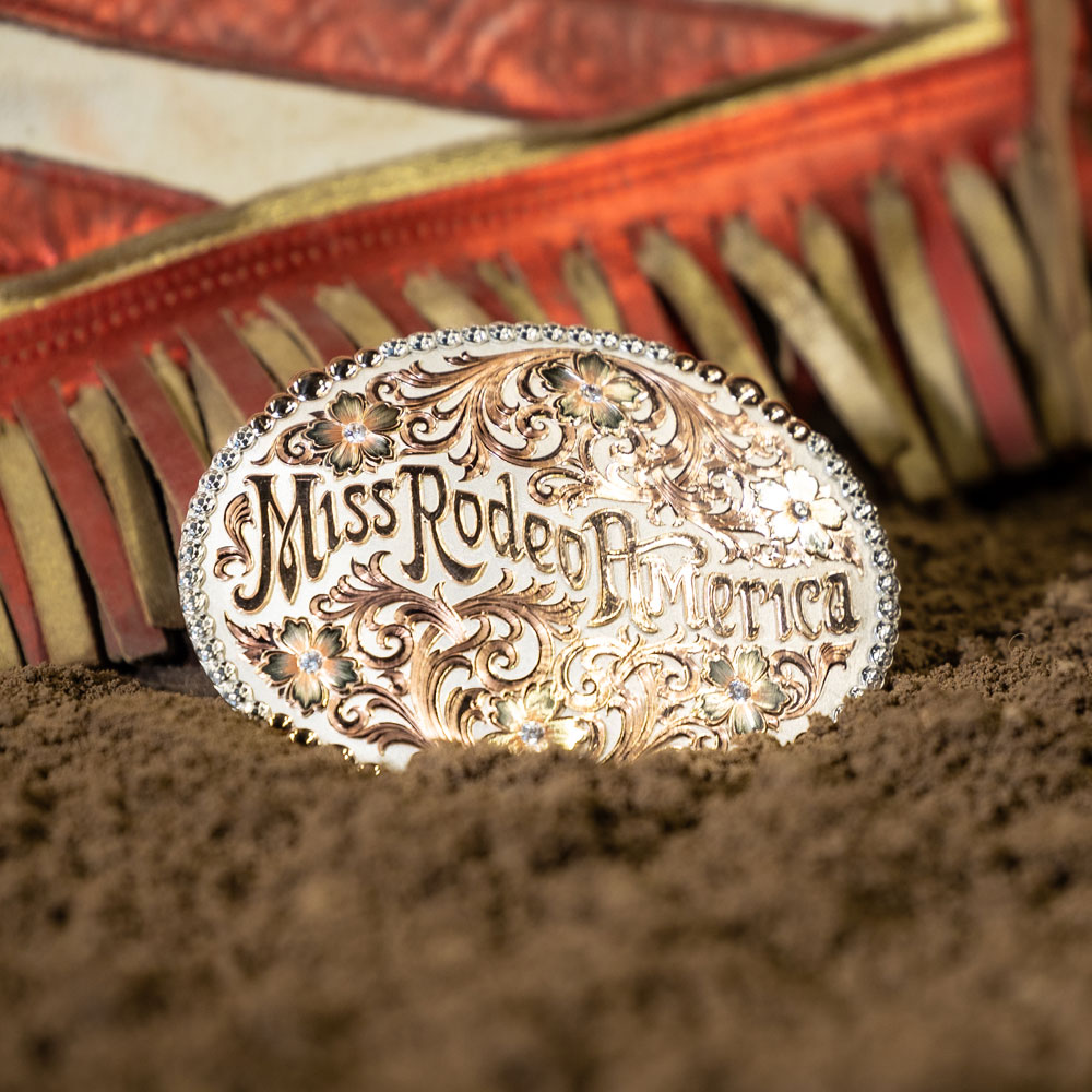 The silver and gold Miss Rodeo America buckle posed in arena dirt next to Miss Rodeo America chaps.